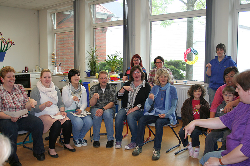 Auf dem Foto sehen Sie die Kursteilnehmer in der Runde sitzen und eine Handbewegung der Gebärdensprache ausführen.