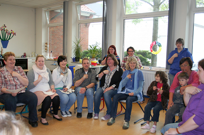 Auf dem Foto sehen Sie die Kursteilnehmer in der Runde sitzen und eine Handbewegung der Gebärdensprache ausführen.